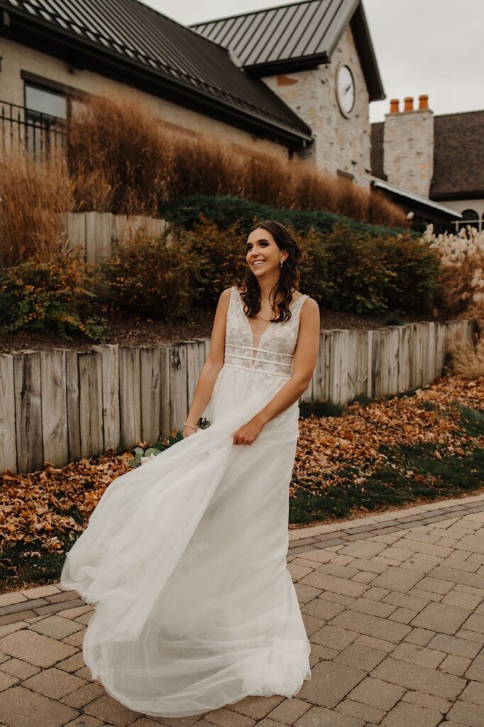 A bride holds her dress down from the wind and smiles while standing on a path next to a building with fall colored leaves on the ground behind her