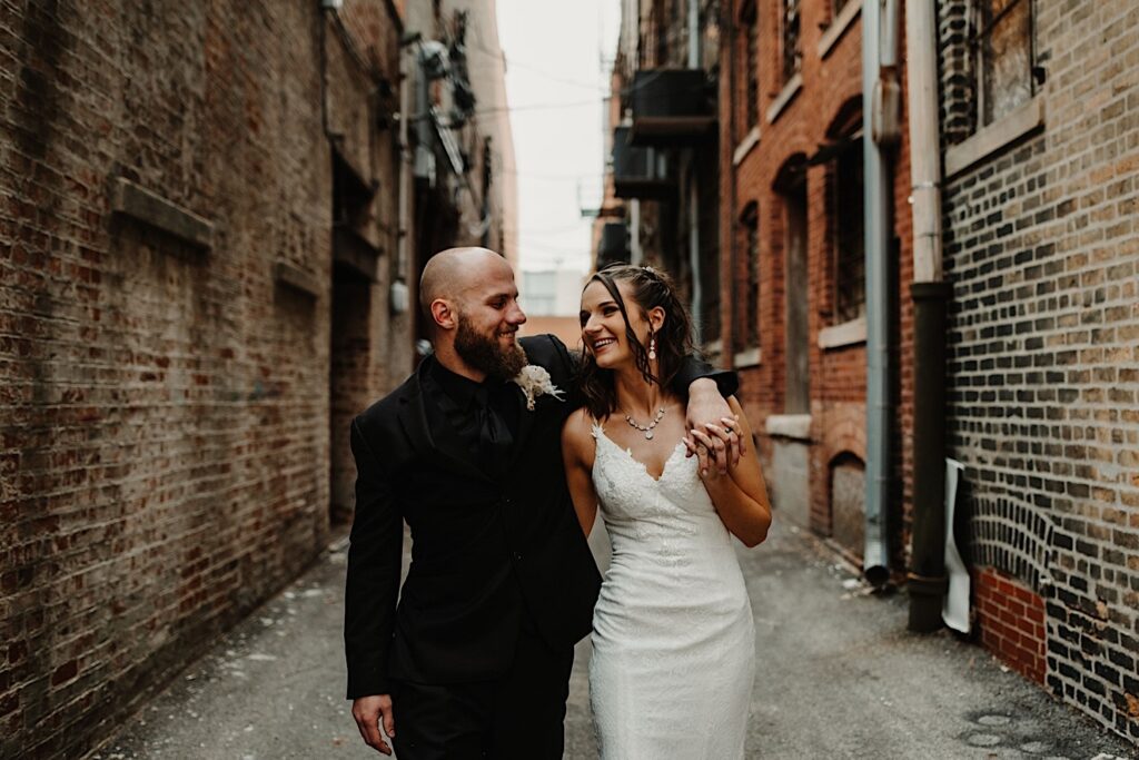 A bride and groom smile at one another while the groom wraps his arm around the brides shoulder while they walk down an alley outside their wedding venue Reality on Monroe