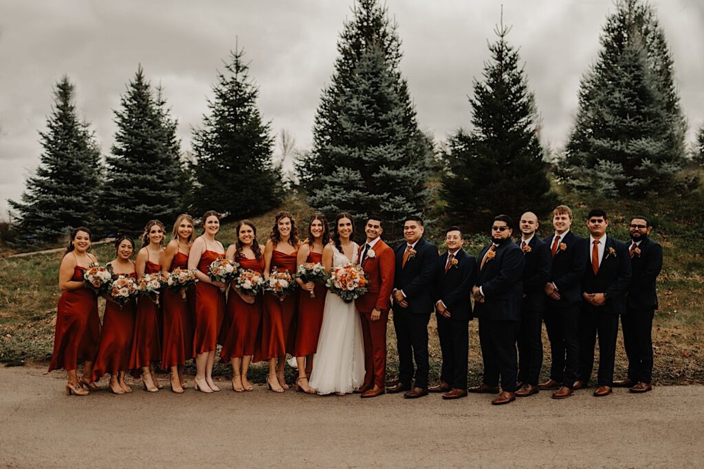 A bride and groom stand in the middle of their wedding parties as they all smile at the camera while outside their wedding venue Mistwood Golf Club in the fall