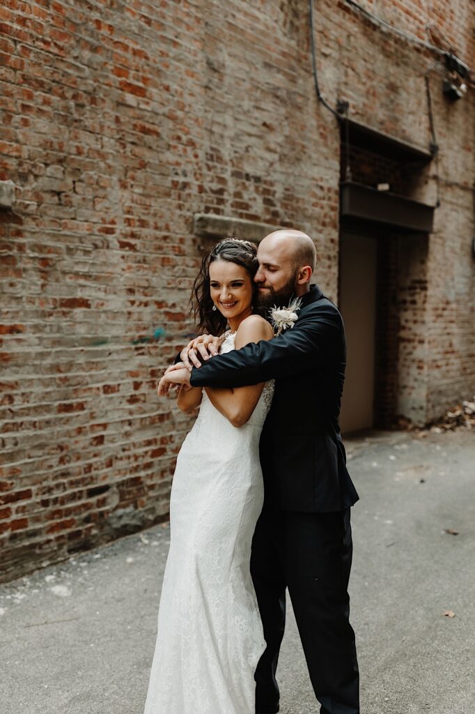 A bride smiles as the  groom stands behind her and hugs her as they stand in an alley in front of a brick wall
