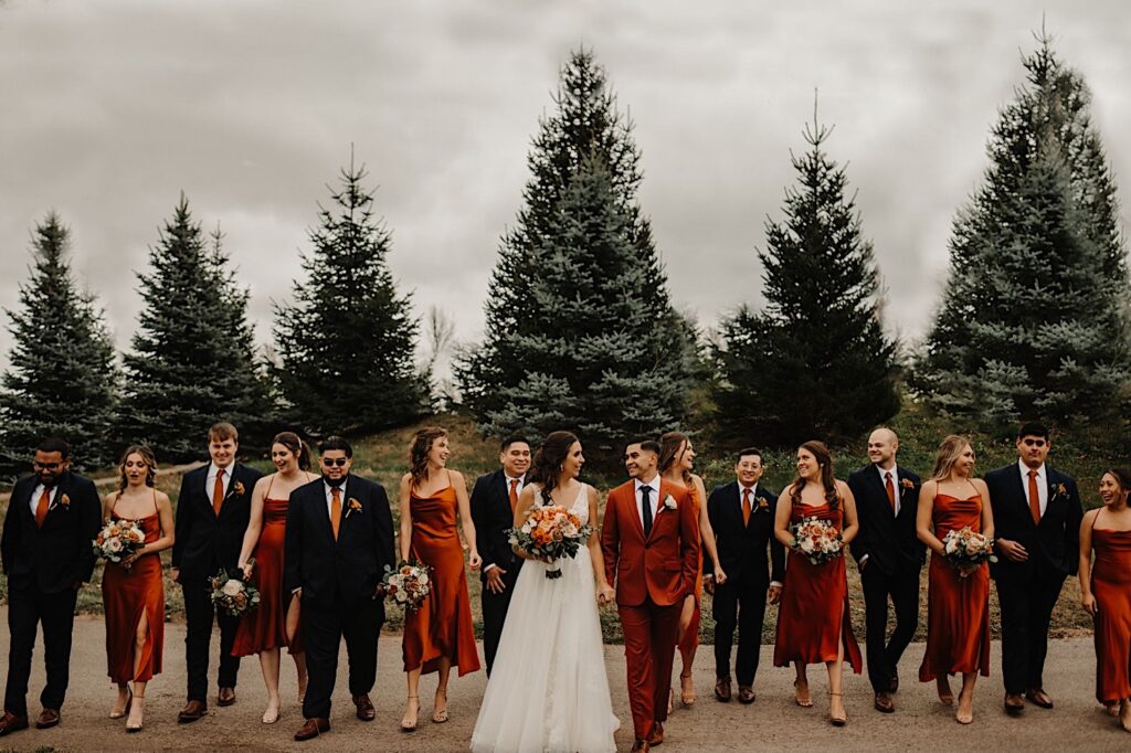 A bride and groom hold hands and look at one another while smiling at the camera with their wedding parties on either side of them walking along and talking with one another while on a path outside their wedding venue Mistwood Golf Club in the fall