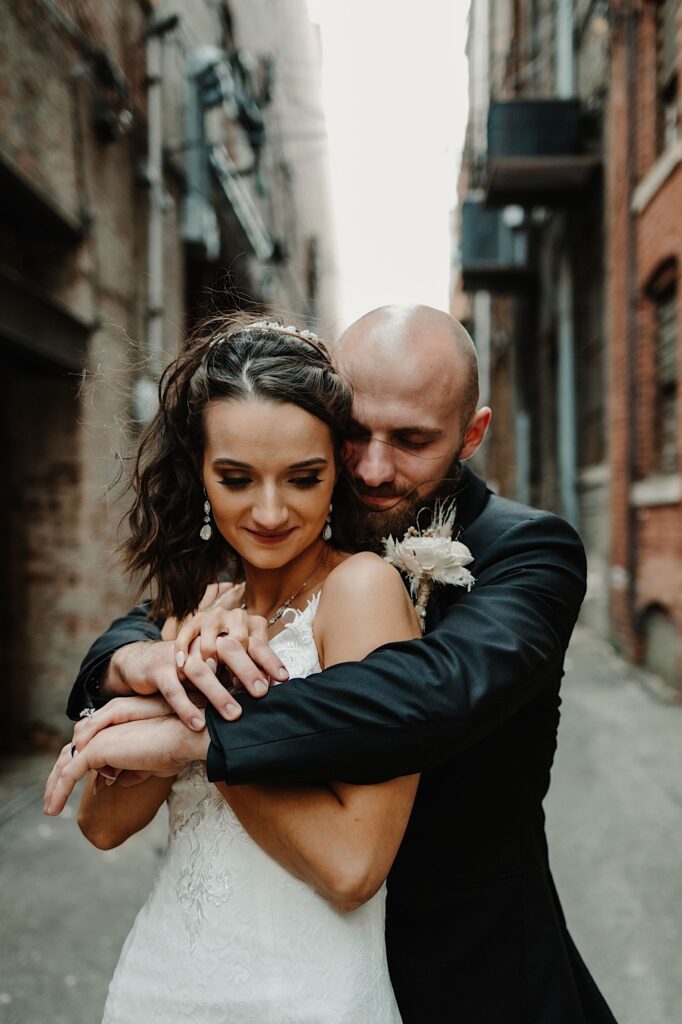 A bride smiles as the  groom stands behind her and hugs her as they stand in an alley in front of a brick wall