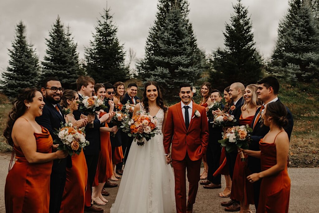 A bride and groom hold hands and smile at the camera while walking towards it on a path outside their wedding venue Mistwood Golf Club in the fall, on either side of them are their wedding parties clapping and cheering for them