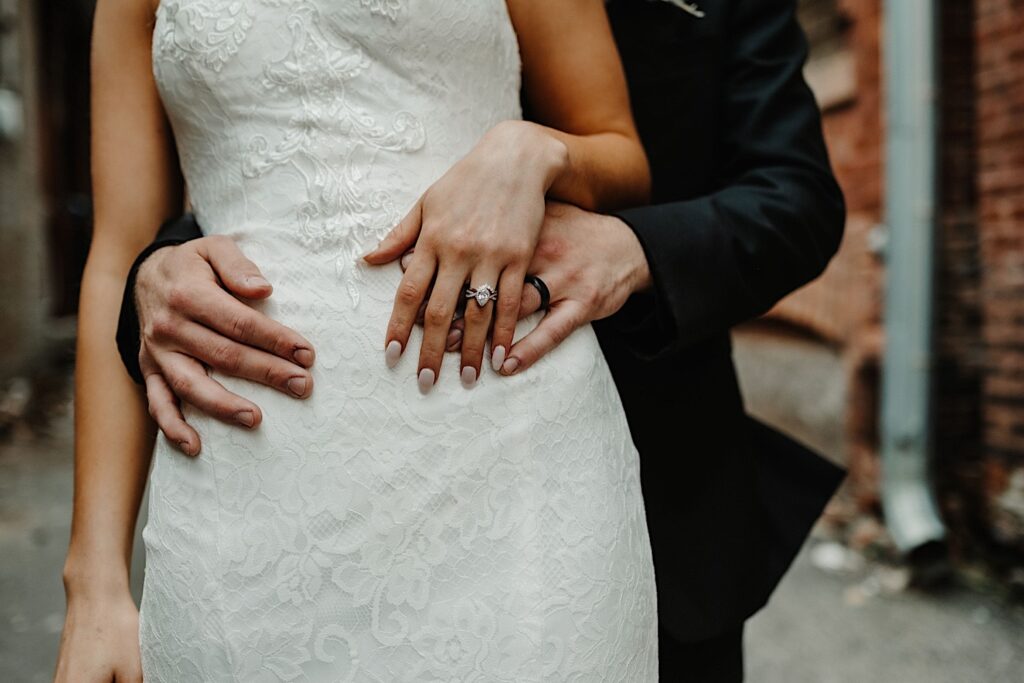 Chest down photo of a bride standing in front of a groom with his hands on her waist and her hands over his showing off both of their wedding rings