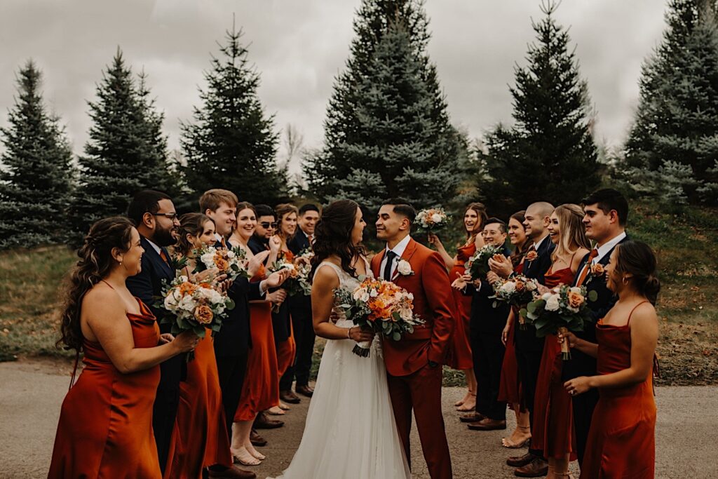 A bride and groom face one another and are about to kiss while on a path outside their wedding venue Mistwood Golf Club in the fall, on either side of them are their wedding parties who cheer and clap for them