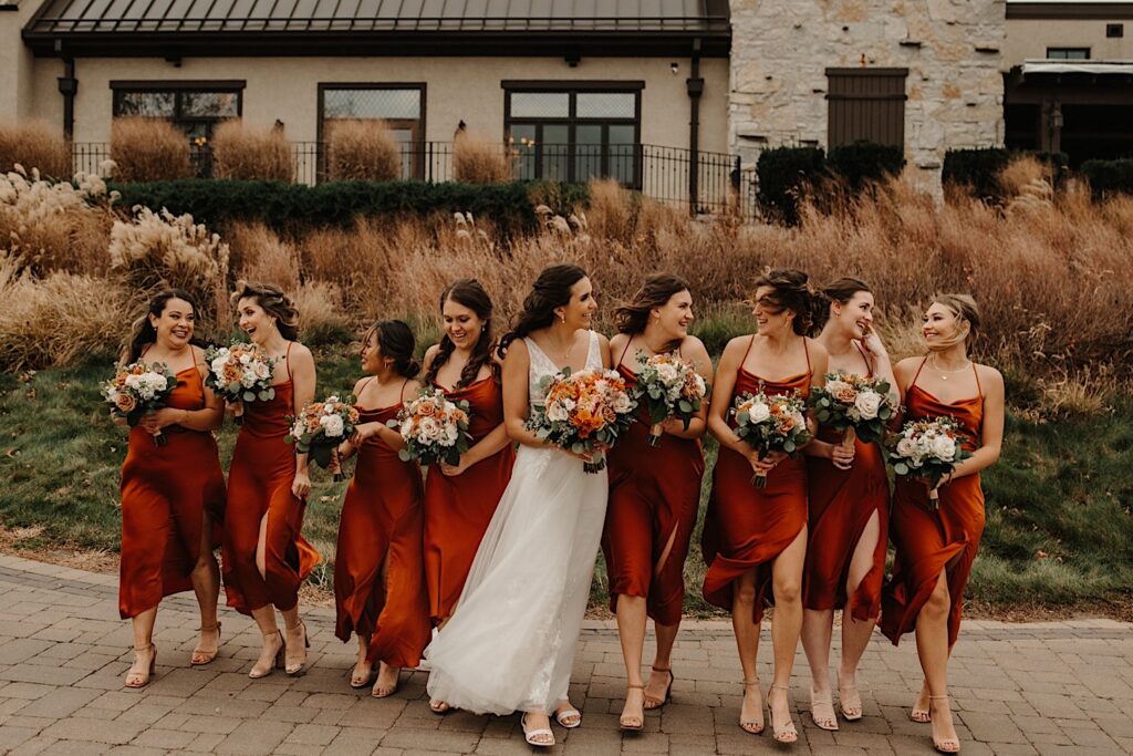 A bride walks on a path and smiles with her bridesmaids on either side of her while outside their wedding venue Mistwood Golf Club in the fall