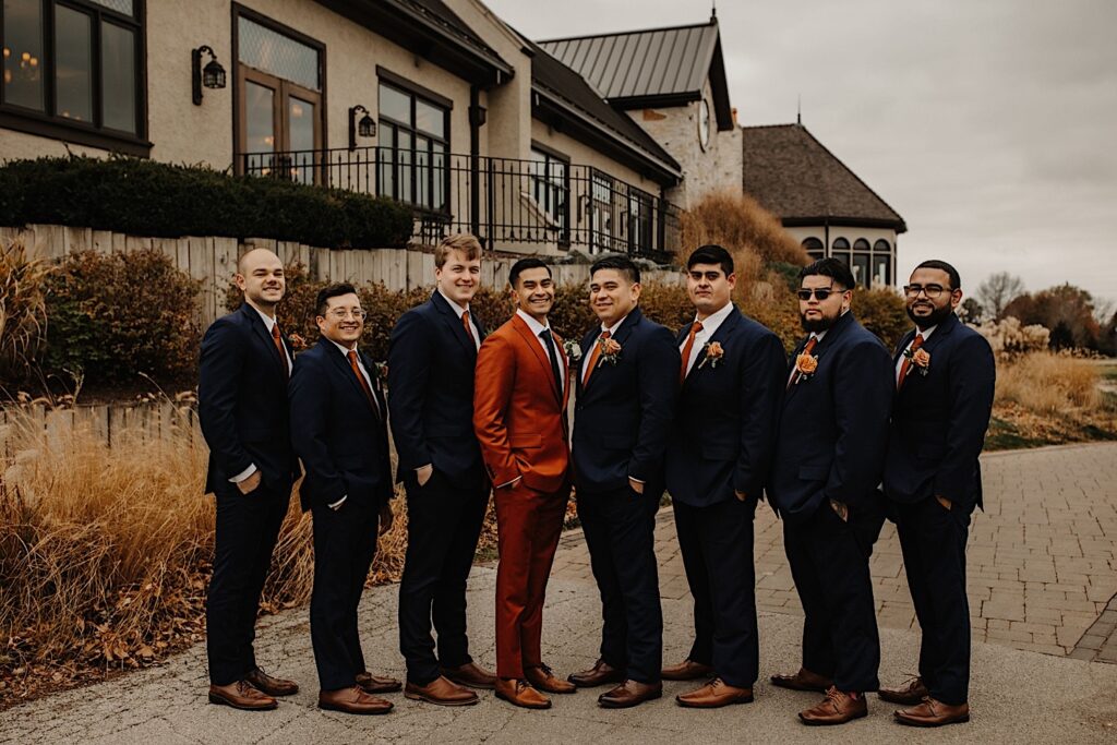 A groom stands on a path with his groomsmen standing on either side of him as they all smile at the camera while outside their wedding venue Mistwood Golf Club in the fall