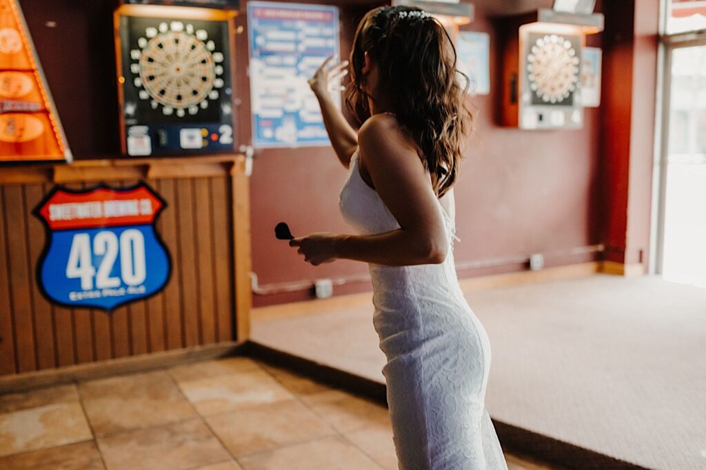 A bride facing away from the camera throws a dart at a dartboard in a bar