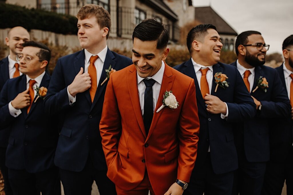 A groom laughs while looking down as his groomsmen laugh and smile on either side of him outside their wedding venue Mistwood Golf Club in the fall