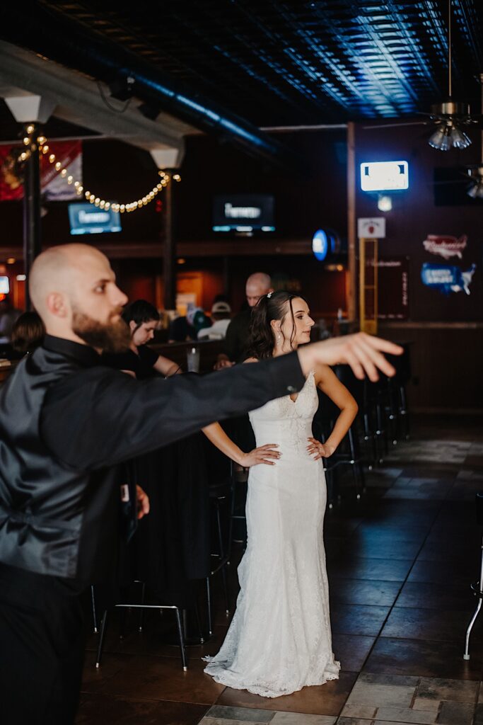 A groom throws a dart while the bride stands behind him watching with her hands on her hips as they hang out in a bar