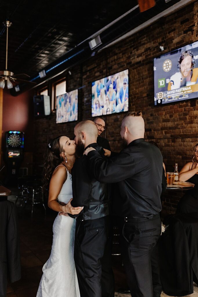 A bride and groom in a bar kiss one another while a groomsmen massages the groom's shoulders