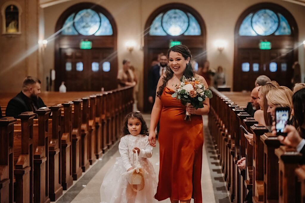 A flower girl walks alongside a smiling bridesmaid down the aisle of a church during a wedding ceremony