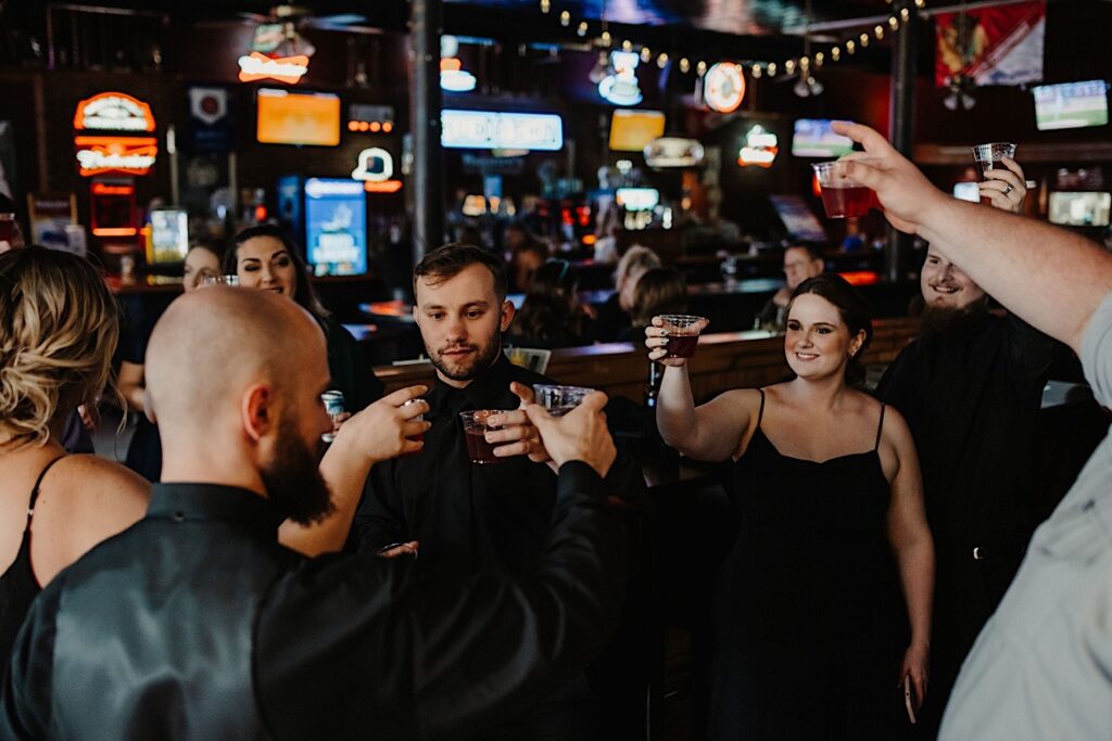 A groom raises a glass with the members of the wedding party around him all raising their glasses as well as they hang out in a bar