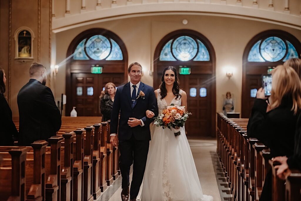 A bride smiles while walking with her father down the aisle of a church for her wedding ceremony