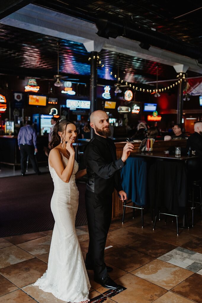 A bride and groom stand side by side each with a dart in their hand while at a bar and are about to throw them at the dart board