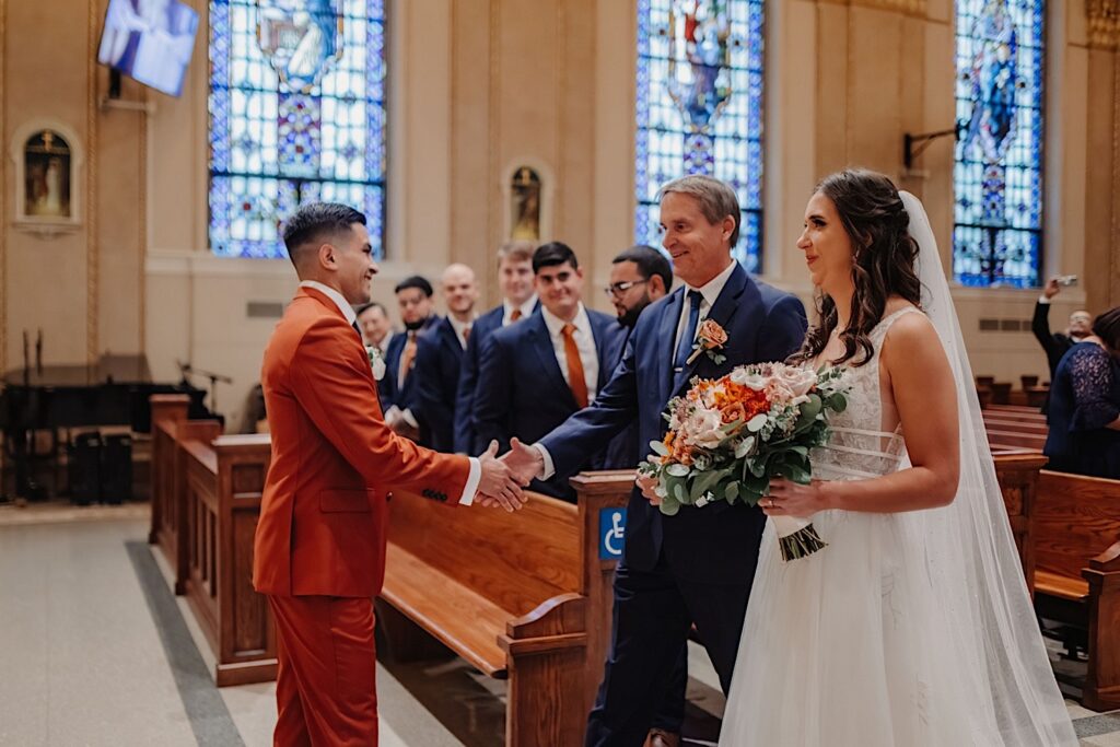 A groom smiles as he shakes the hand of the father of the bride as he walks his daughter down the aisle of the church for their wedding ceremony