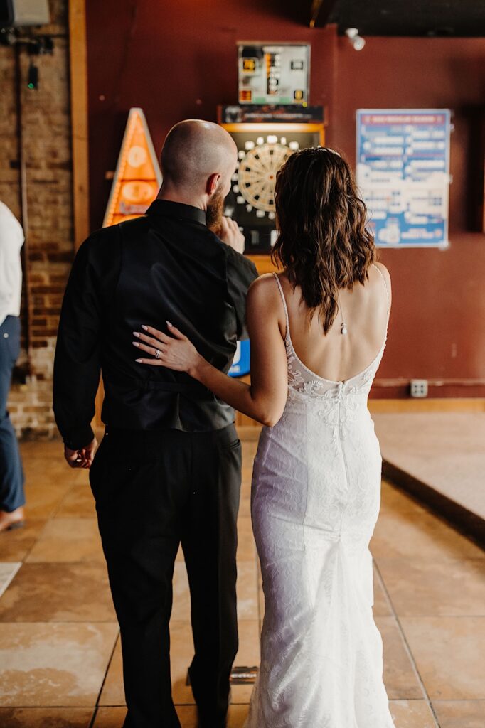 A bride and groom face away from the camera towards a dartboard in a bar as the groom is about to throw a dart and the bride has a hand on his back