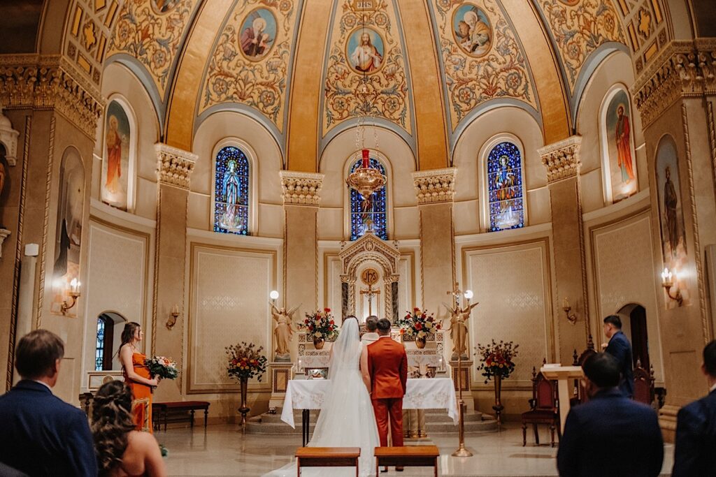 A bride and groom stand in the center of a church during their wedding ceremony facing away from the camera