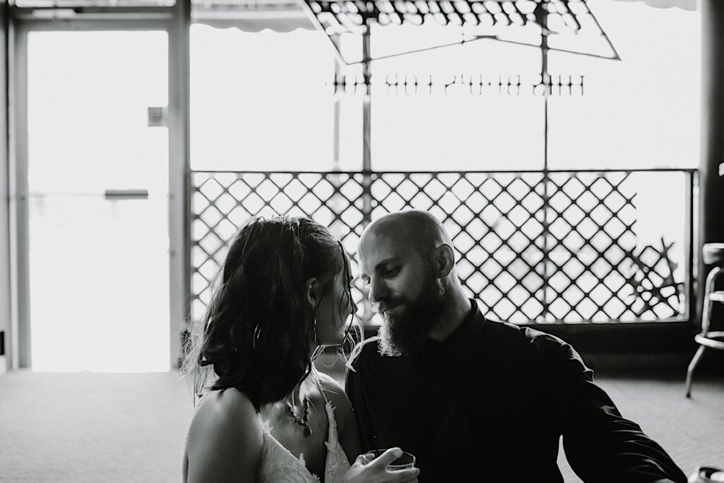 Black and white photo of a bride and groom sitting with one another inside a bar about to kiss one another