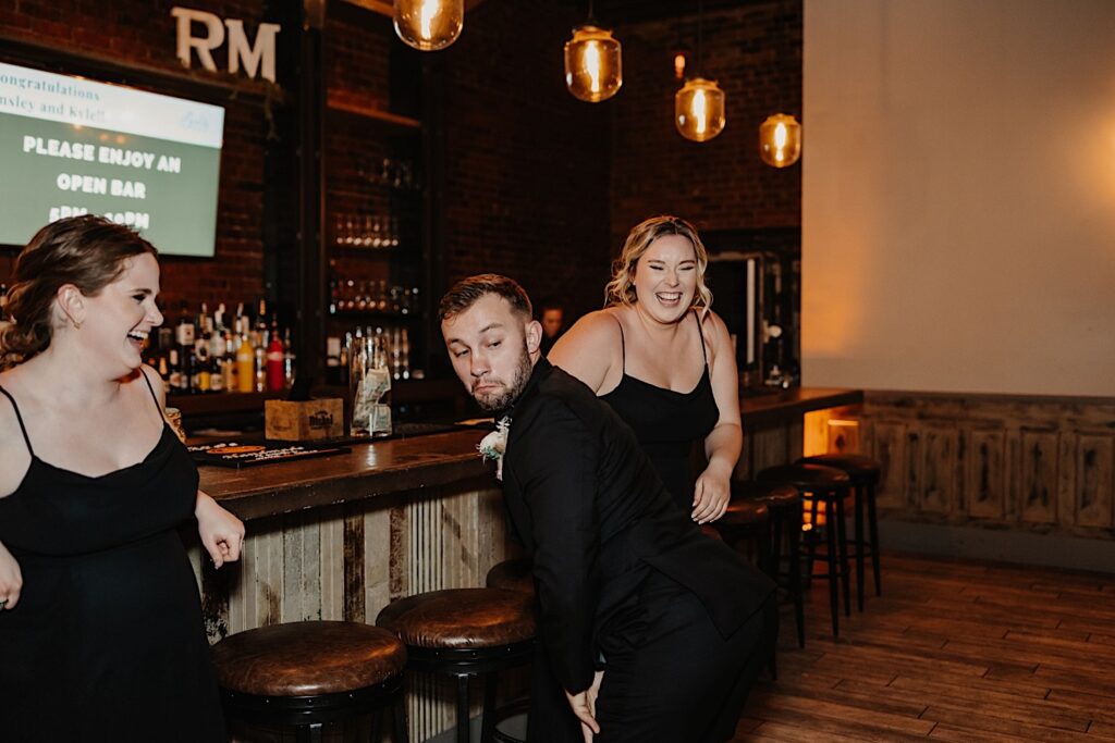 A groomsman twerks at the bar and makes a funny face while two bridesmaids laugh on either side of him during a wedding reception at Reality on Monroe