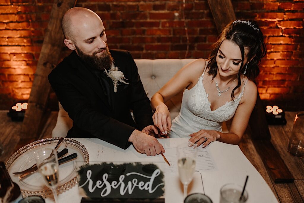 A bride and groom sitting at their head table in front of a brick wall inside Reality on Monroe smile as they sign their wedding paperwork