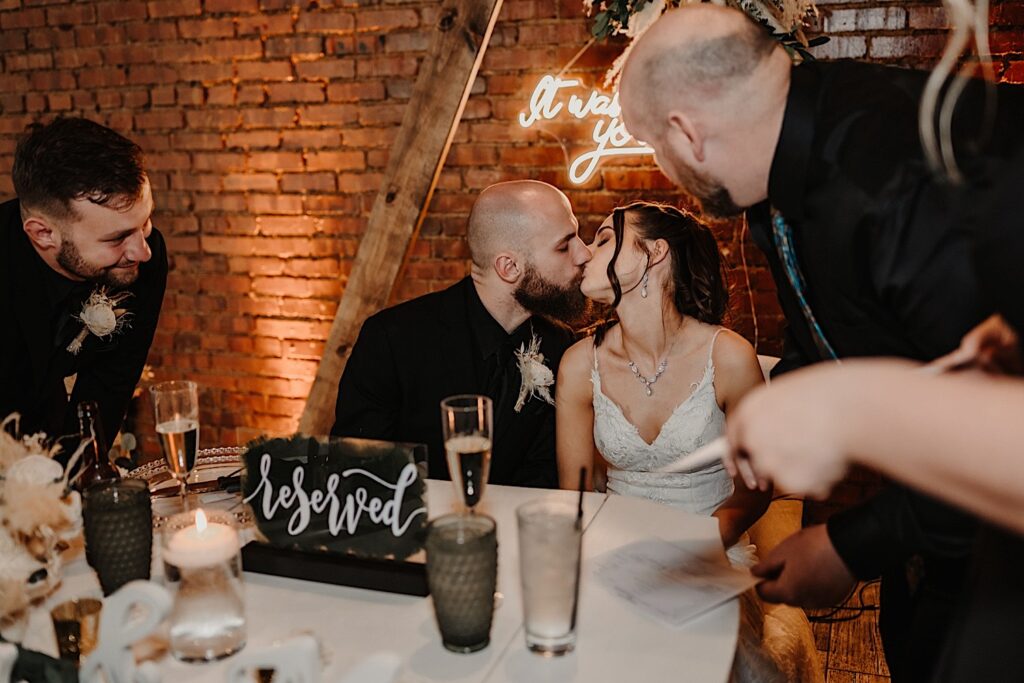 A bride and groom sitting at their head table in front of a brick wall inside Reality on Monroe kiss one another while guests watch after they sign their wedding paperwork