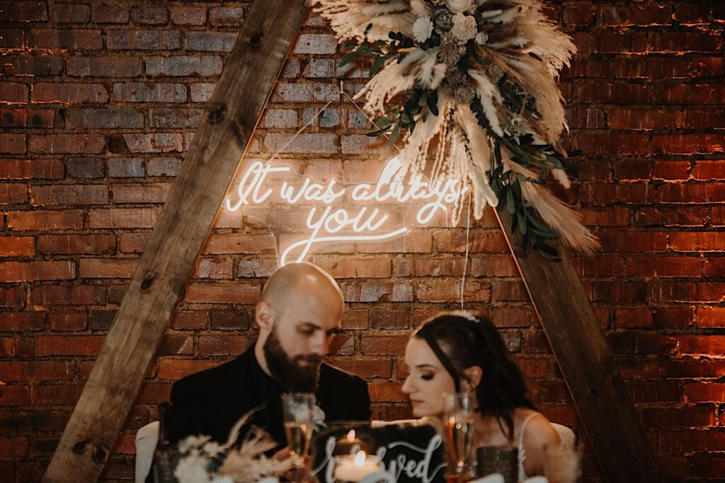 A bride and groom sit out of focus at their head table in front of a brick wall and a neon sign that reads "It was always you" inside their wedding venue Reality on Monroe