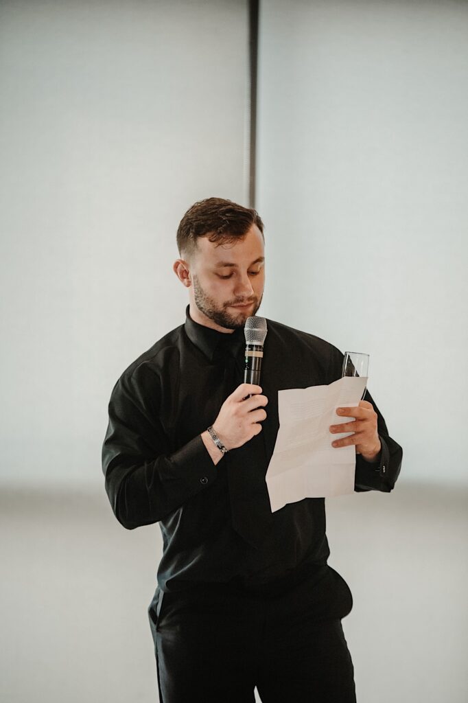 A groomsman stand with a microphone, piece of paper, and champagne glass in hand while giving a speech during a wedding reception