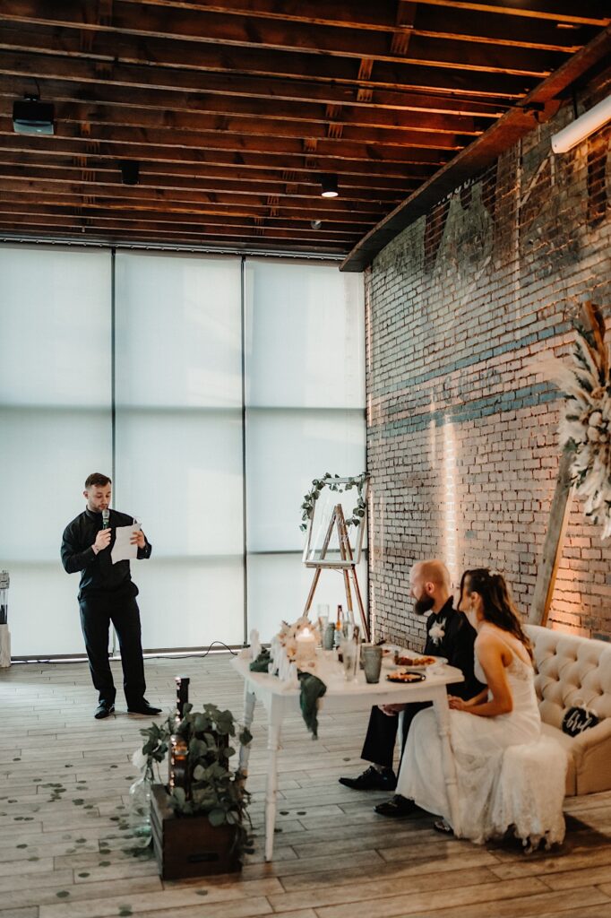 A bride and groom seated at their head table look to their right towards a groomsman standing while giving a speech to them