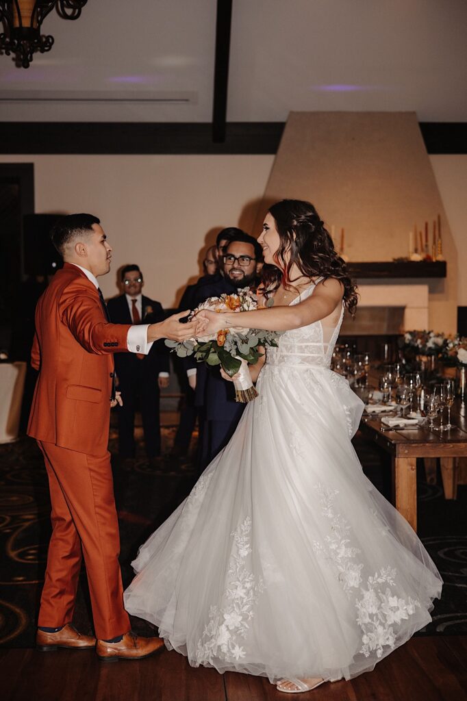 A bride and groom share their first dance together next to their table as groomsmen behind them watch during their indoor reception