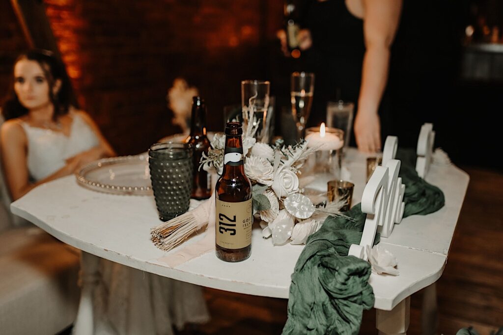 A bottle of 312 beer sits on a white wooden table decorated for a wedding with the bride out of focus in the background during a wedding reception at Reality on Monroe