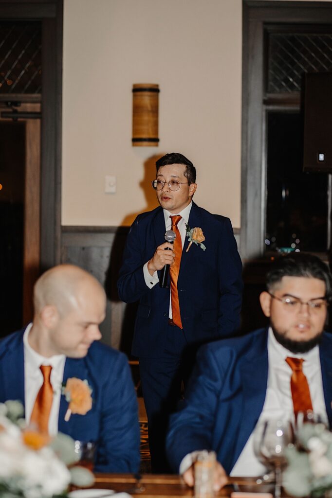 A man stands with a microphone in front of a table of groomsmen and gives a speech during an indoor wedding reception
