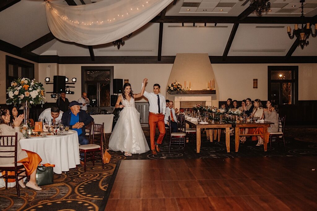 A bride and groom hold hands as they walk out to the dance floor for their first dance as guests watch during their indoor fall wedding reception at Mistwood Golf Club