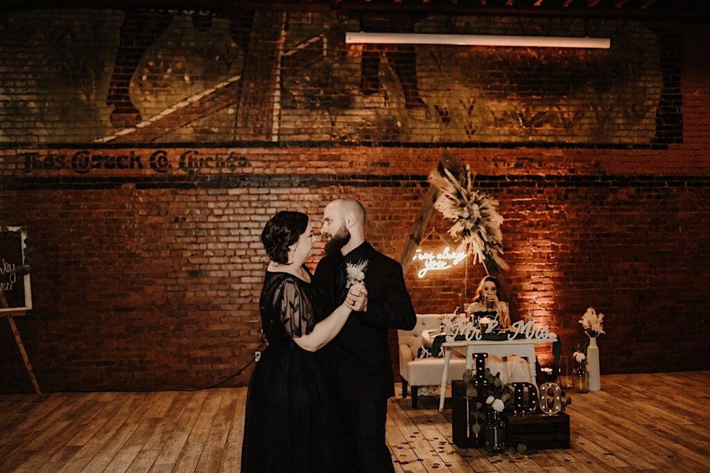 A groom dances with his mother during a wedding reception at Reality on Monroe, behind them is the bride sitting at the head table with a drink and a brick wall of the building