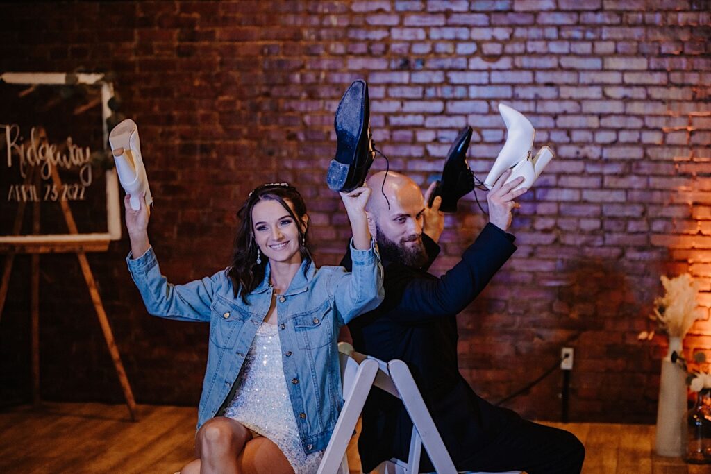 A bride and groom seated back to back each have one of each other's shoes in their hands raised in the air for the shoe game during their wedding reception at Reality on Monroe