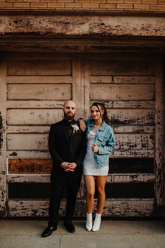 A bride and groom stand next to one another and pose in front of an old wooden double door while smiling at the camera