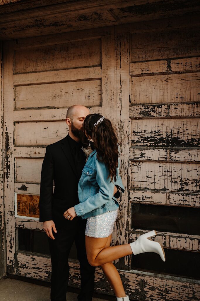 A bride kicks her leg up behind her while kissing the groom standing next to her while they stand in front of an old wood double door