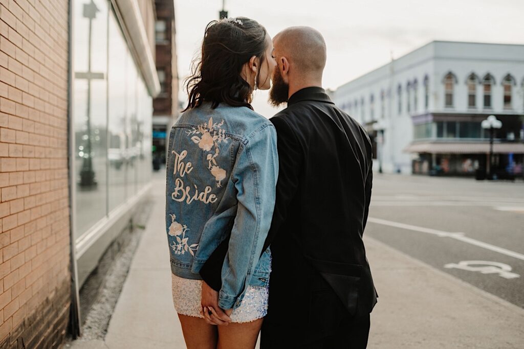 A bride wearing a jean jacket that reads "The Bride" on it holds the grooms hand as the two walk away from the camera down a sidewalk in Bloomington Illinois