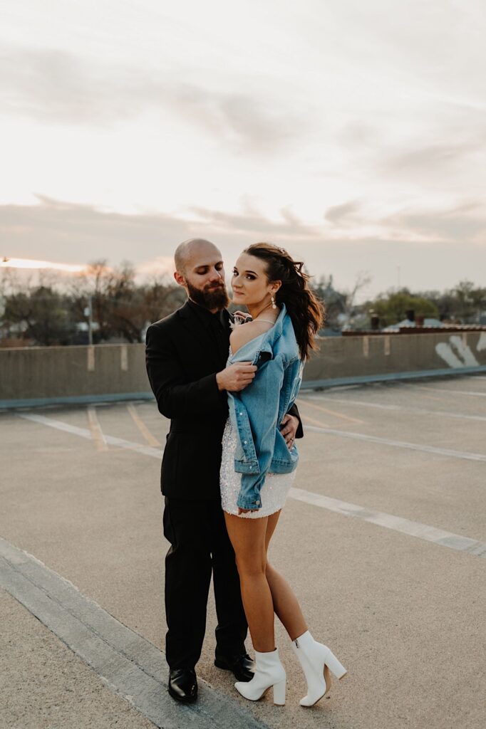 A bride looks over her shoulder back at the camera while the groom pulls the jean jacket she's wearing off her should while the two stand atop a parking garage
