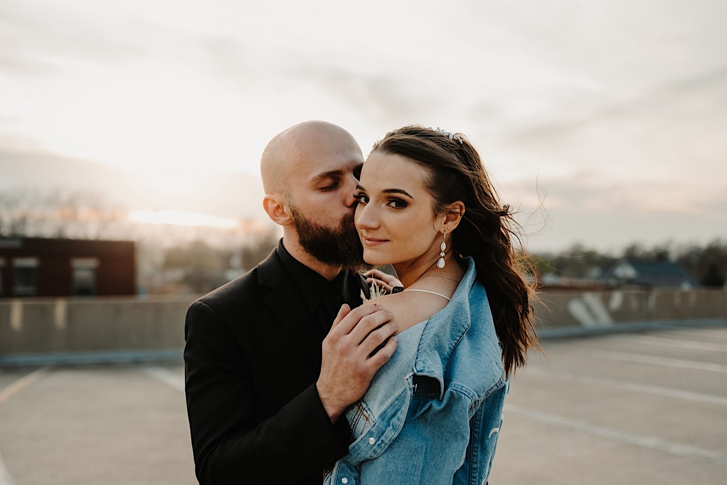 A bride smiles over her shoulder at the camera while the groom kisses her cheek as the sun sets behind them while they stand atop a parking garage