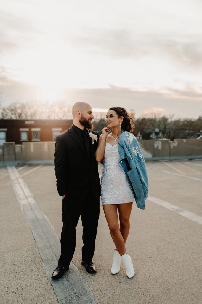 A bride and groom stand side by side and smile at one another while standing atop a parking garage as the sun sets behind them, the bride has a jean jacket over her shoulder
