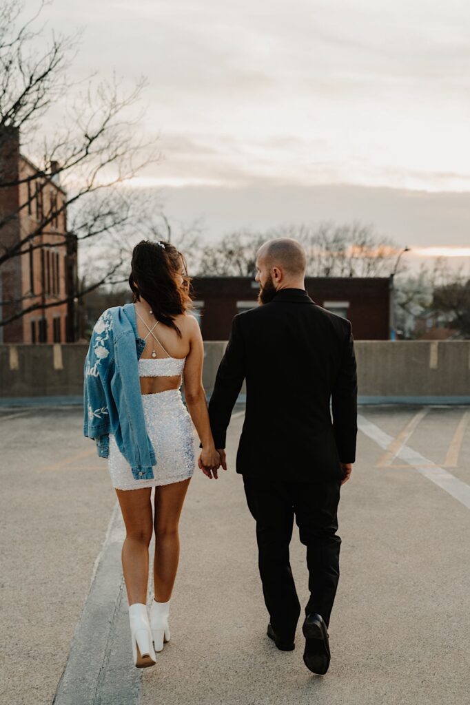 A bride and groom walk hand in hand away from the camera atop a parking garage during sunset while looking at one another, the bride has a jean jacket over her shoulder
