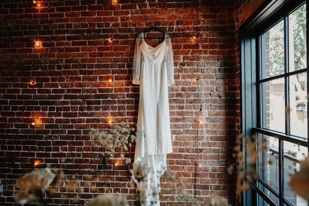 A wedding dress hangs on a brick wall next to a window with string lights hanging next to the dress