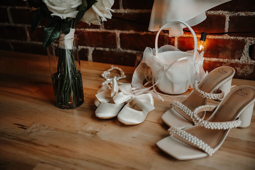 Wedding shoes, a flower vase, a white basket, and a pearl necklace lay on a wood table next to a brick wall
