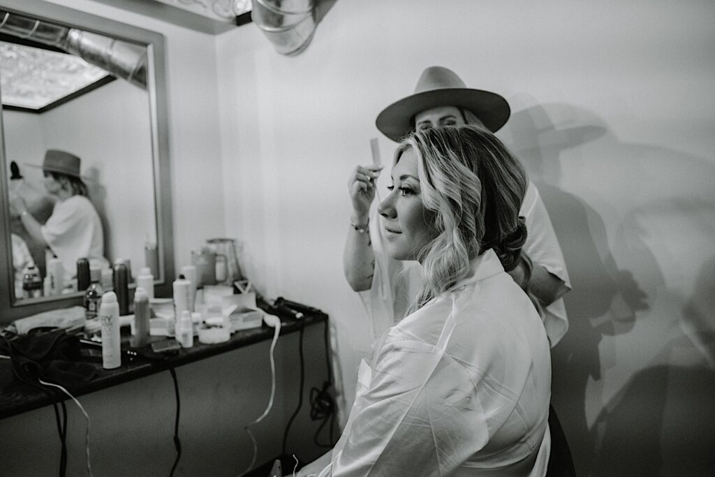 Black and white photo of a bride sitting in front of a mirror as a woman puts her makeup on before her wedding day
