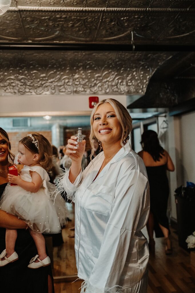 A bride smiles at the camera while holding a drink before getting ready for her wedding day with bridesmaids in the background