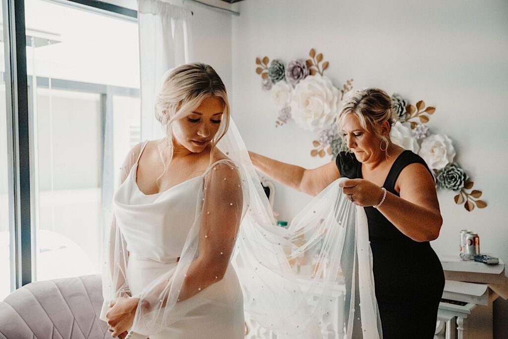 A bride in her wedding dress looks over her shoulder while her mother behind her adjusts her veil while getting ready for her wedding day at Venue CU in Champaign