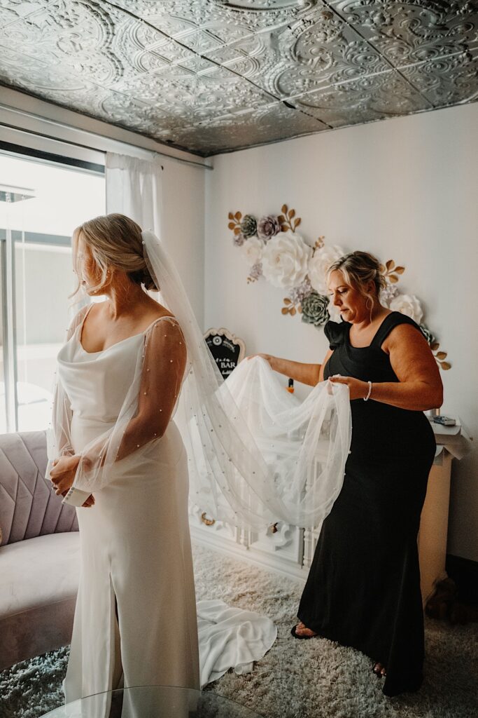 A bride in her wedding dress looks out a window to her right as her mother behind her adjusts her veil