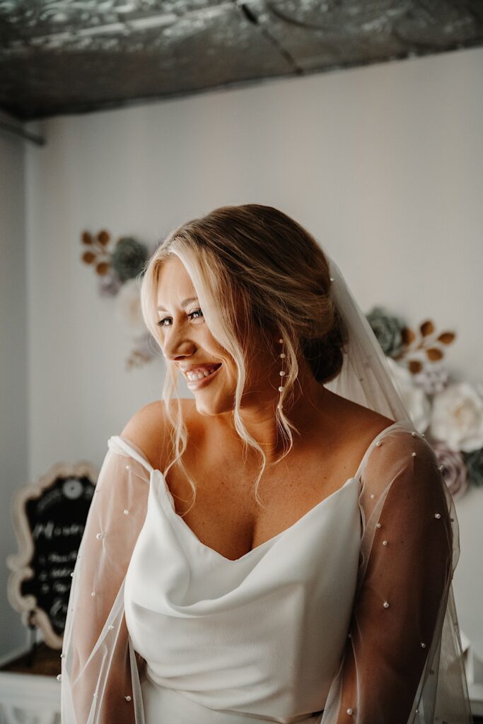 A bride smiles while in a room wearing her wedding dress while getting ready for her wedding day