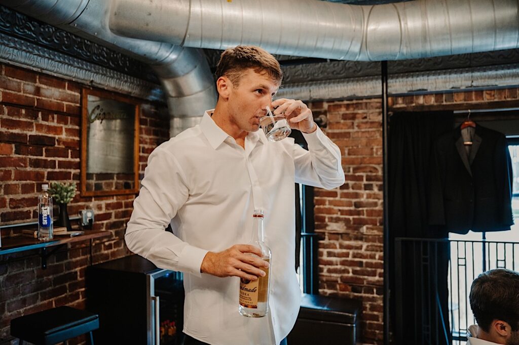 A man holding a bottle of vodka drinks from a glass while in a brick room getting ready for a wedding day
